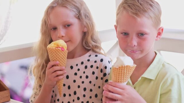 Girl and boy eat and licking ice cream in cone. prescholler friends enthusiastically eating colourful ice cream in cone. The fair-haired brother and sister bought sweet ice cream in cone at a cafe.4K