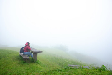 Asian young woman in red raincoat and bag sitting on wooden bench in green grass field and foggy with mist on mountain in rainy season. Travel in Asian