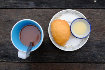 Yang fried dumpling or Bread fried bun recipe with sweetened condensed milk in a bowl and a cup of hot chocolate on wooden table in tropical garden. Asain Breakfast in Thailand