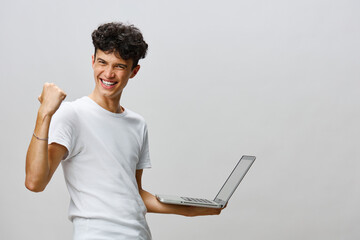 Happy young man celebrating success while holding a laptop, dressed in a casual white t shirt on a neutral grey background Expression of joy and achievement captured in this modern setting