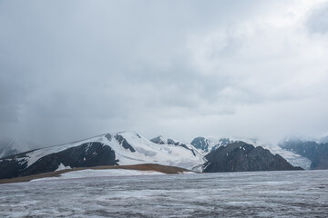 Dramatic vast misty top view from ice pass to big glacier tongue among sharp rocks and large snow-capped mountains in rainy low clouds. Dark atmospheric mountain silhouettes in rain in gray cloudy sky