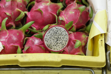 Dragon fruit cut in half to show the flesh inside is placed on a basket. Dragon fruits are piled up in a yellow basket.