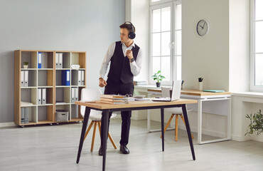 Young businessman or corporate employee having fun during a break at work. Happy business man in headphones standing by his desk in a modern office interior, listening to cool music and dancing