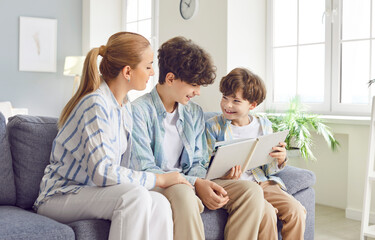 Portrait of a happy smiling young mother parent looking through a family photo album sitting on sofa at home with her children sons. Kids enjoying leisure time with mom. Family good memories