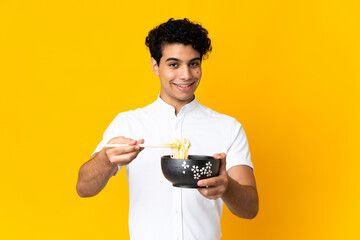 Young Venezuelan man isolated on yellow background holding a bowl of noodles with chopsticks and offering it