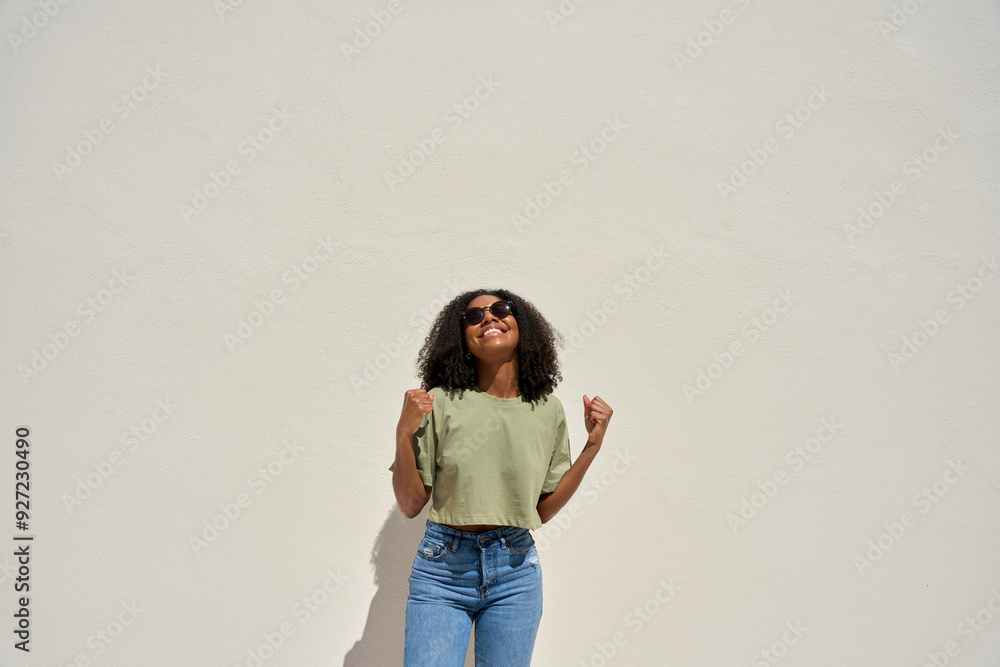 Wall mural Happy young African American woman winner feeling joy celebrating win victory standing at white wall background. Funky gen z Afro Black girl dancing having fun at sunny summer party. Copy space.