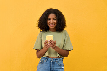 Young happy African gen z teen girl standing at yellow wall looking at camera holding smartphone....