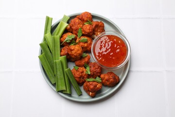 Baked cauliflower buffalo wings with celery and sauce on white tiled table, top view
