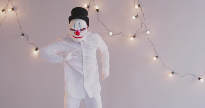 Dancing child in scary Halloween costume. Kid dressed as creepy clown having fun at Halloween party. Boy in white shirt, black tophat and evil clown mask with red nose dancing on light gray background