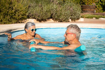 Elderly couple enjoying a sunny day in the pool, exercising with foam noodles while smiling and having fun