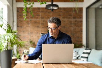 Busy mature middle aged professional businessman executive using laptop at work writing notes. 50...