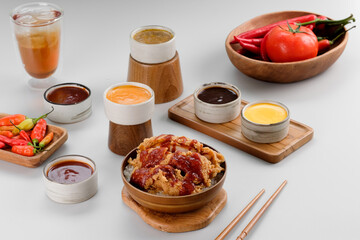 A rice bowl topped with crispy chicken and drizzled with spicy red sauce, accompanied by a variety of sauces around it.