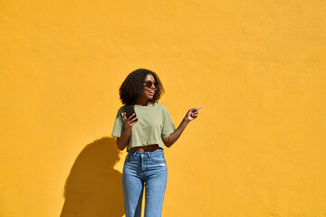 Young happy African American girl wearing sunglasses standing at yellow wall pointing at copy space holding smartphone. Smiling Black woman using mobile cell phone advertising ecommerce summer sale.