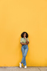 Young happy cheerful pretty African American gen z teen girl standing at yellow wall. Smiling Black woman looking at camera presenting summer promotion posing on color background. Vertical full body. © insta_photos