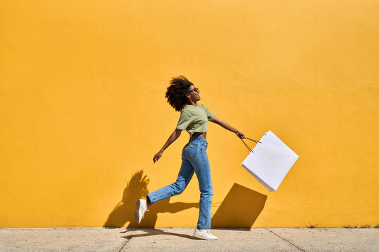 Happy Black girl customer walking at yellow wall holding shopping bag. Cheerful young African American woman shopper wearing sunglasses jumping buying making summer fashion sale purchase. Full body.