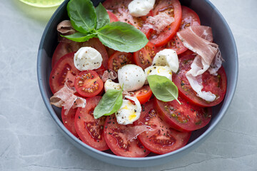 Italian caprese salad served with torn prosciutto, horizontal shot on a grey granite surface, middle close-up, selective focus