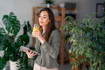 Serene Caucasian lady sips orange juice while browsing on a digital tablet, clad in casual work...