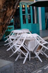 A photo of a deserted outdoor cafe with white chairs and a table piled on top of each other. The cafe has a teal-colored door and awning, and the background shows a tree and a building. 