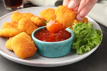 Woman dipping nugget into chili sauce at grey table, closeup