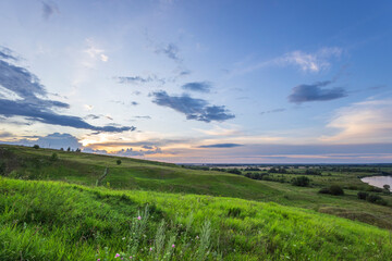 A panoramic view of a lush, green landscape at sunset, showcasing rolling hills, a winding river,...