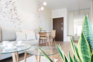 Modern living room with dining room table with stylish chairs. White wall with wallpaper and door in luxurious interior in apartment with plant. Hotel with furniture.	
