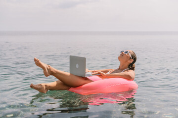 A woman is floating on a pink inflatable raft in the ocean while holding a laptop. Concept of...