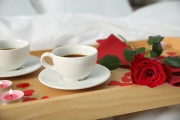 Wooden tray with burning candles, rose, cups of coffee and red paper hearts on bed indoors, closeup