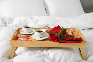 Wooden tray with burning candles, rose, cups of coffee and red paper hearts on bed indoors