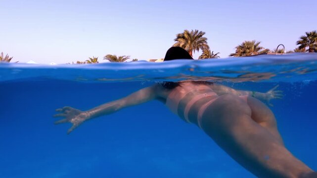 A young woman swims gracefully in a pool with a backdrop of palm trees. Perfect for capturing relaxation, luxury, and the essence of a tropical getaway.