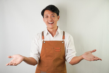 Excited young asian man wearing rolled-up white sleeve shirt and brown apron is opening hands receiving something with smiling face expression. Isolated over white background