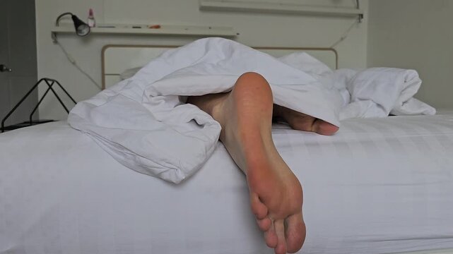 Close-up of bare feet under a white blanket on a bed. Bare feet sticking out from under a white blanket and rubbing leg against leg.