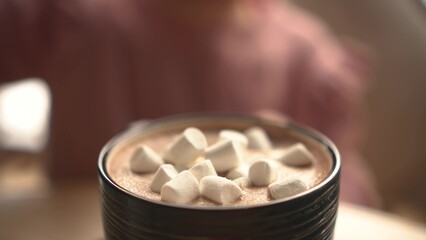 Girl sprinkling marshmallows into a cup of hot chocolate on a green saucer, placed on a wooden table, with a cozy cafe background. Close-up.