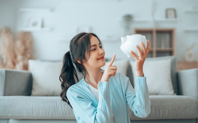 Young Asian woman holding piggy bank and sitting on the floor at home. Save money and financial...