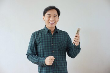 Adult Asian man in green long sleeve flannel shirt looking and smiling at camera while left hand holding smartphone and right hand making fist. Happy, cheerful and satisfaction expression.