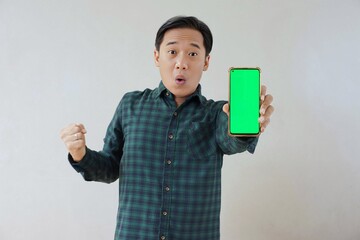 Adult Asian man in green long sleeve flannel shirt holding and showing smartphone display with green screen to camera with left hand while right hand making fist. Happy and cheerful expression