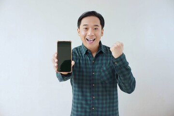 Adult Asian man in green long sleeve flannel shirt holding and showing smartphone display with black screen to camera with right hand while left hand making fist. Happy and cheerful expression.