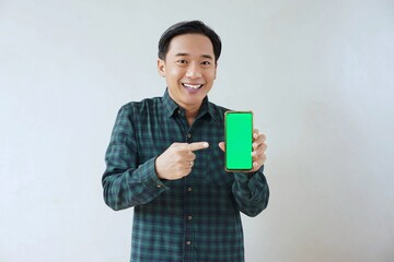 Adult Asian man in green long sleeve flannel shirt showing smartphone display with green screen to camera with left hand while right hand pointing finger to screen. Happy and cheerful expression.