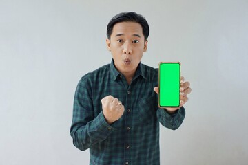 Adult Asian man in green long sleeve flannel shirt holding and showing smartphone display with green screen to camera with left hand while right hand making fist. Happy and cheerful expression