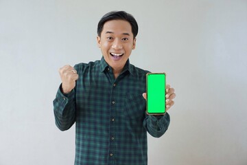 Adult Asian man in green long sleeve flannel shirt holding and showing smartphone display with green screen to camera with left hand while right hand making fist. Happy and cheerful expression.