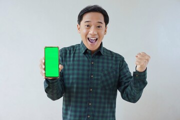 Adult Asian man in green long sleeve flannel shirt holding and showing smartphone display with green screen to camera with right hand while left hand making fist. Happy and cheerful expression.