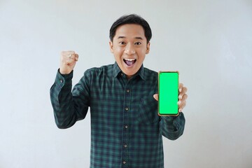 Adult Asian man in green long sleeve flannel shirt holding and showing smartphone display with green screen to camera with left hand while right hand making fist. Happy and cheerful expression.