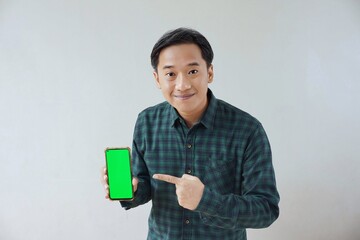 Adult Asian man in green long sleeve flannel shirt showing smartphone display with green screen to camera with right hand while left hand pointing finger to screen. Happy and cheerful expression.