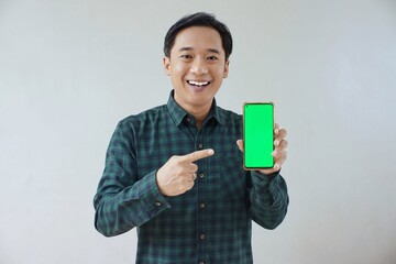 Adult Asian man in green long sleeve flannel shirt showing smartphone display with green screen to camera with left hand while right hand pointing finger to screen. Happy and cheerful expression.