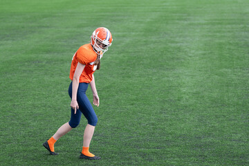 A girl or young woman in an American football uniform stands in the wide receiver position before the run and points her finger downward. In the background, there is green grass on the field.