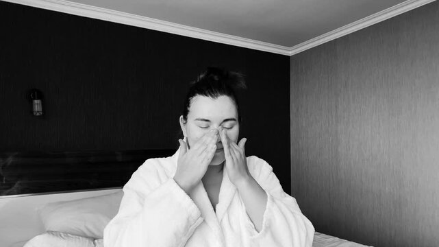 Black and white horizontal 4K video. Young adult woman in bath robe applies moisturizer cream to face in hotel bed room. Concept of skin protection, march 8, relax, body care, mental health, spa, rest