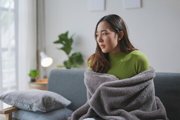 Young asian woman feeling sick and having cold, covering herself with a blanket, while sitting on a sofa at home © amnaj