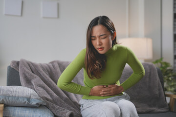 Young woman sits on her couch at home, holding her stomach in pain and discomfort, dealing with...