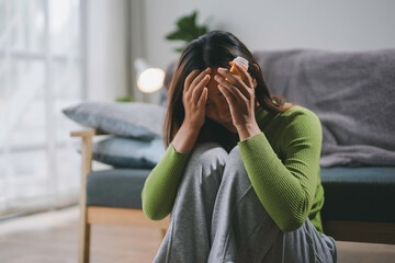 Young woman feeling depressed and holding a pill bottle while sitting on the floor at home