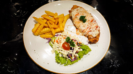 Close-up of Crispy Chicken Steak Topped with Melted Cheese Served with Crinkle Cut Fries and Fresh Salad on a White Plate with a Black Marble Table Background in a Restaurant Setting