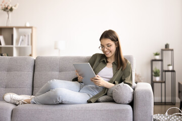 Fototapeta premium Smiling teenager girl in glasses resting seated on couch with tablet device, chatting on-line, web surfing website, search information. Modern wireless tech usage for fun and leisure. Blogging, gen Z